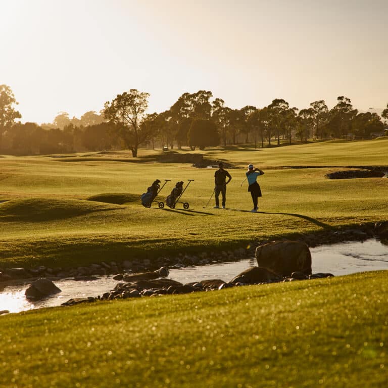A wide, sunlit view of a Tasmanian country club golf course with rolling fairways, scattered trees, and a small creek in the foreground. Two golfers stand near their carts, one mid-swing, framed by warm golden light across the landscape.
